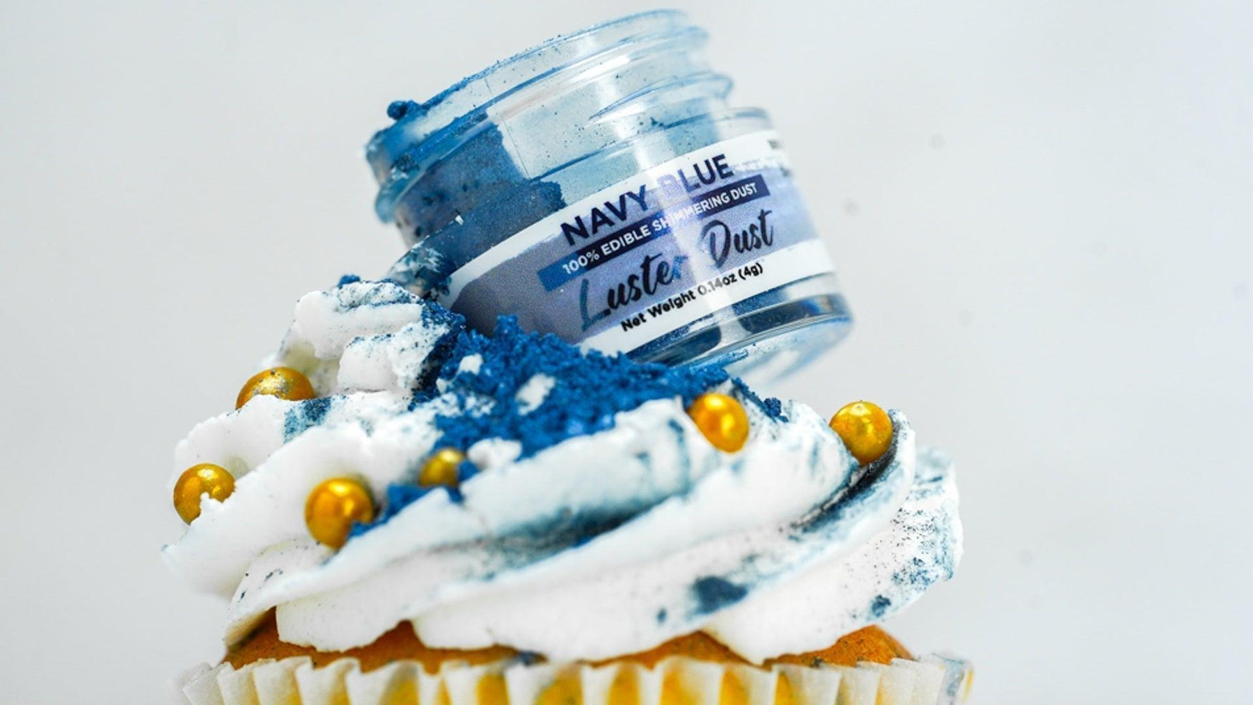 Navy blue edible luster dust spilling from a jar onto a white-frosted cupcake with gold sprinkles.