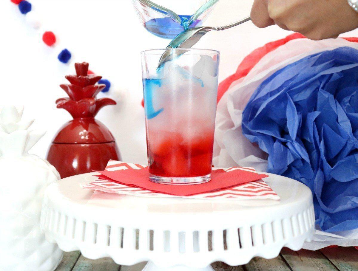 Hand pouring blue liquid into a glass with red liquid and ice, creating a festive red, white, and blue drink.