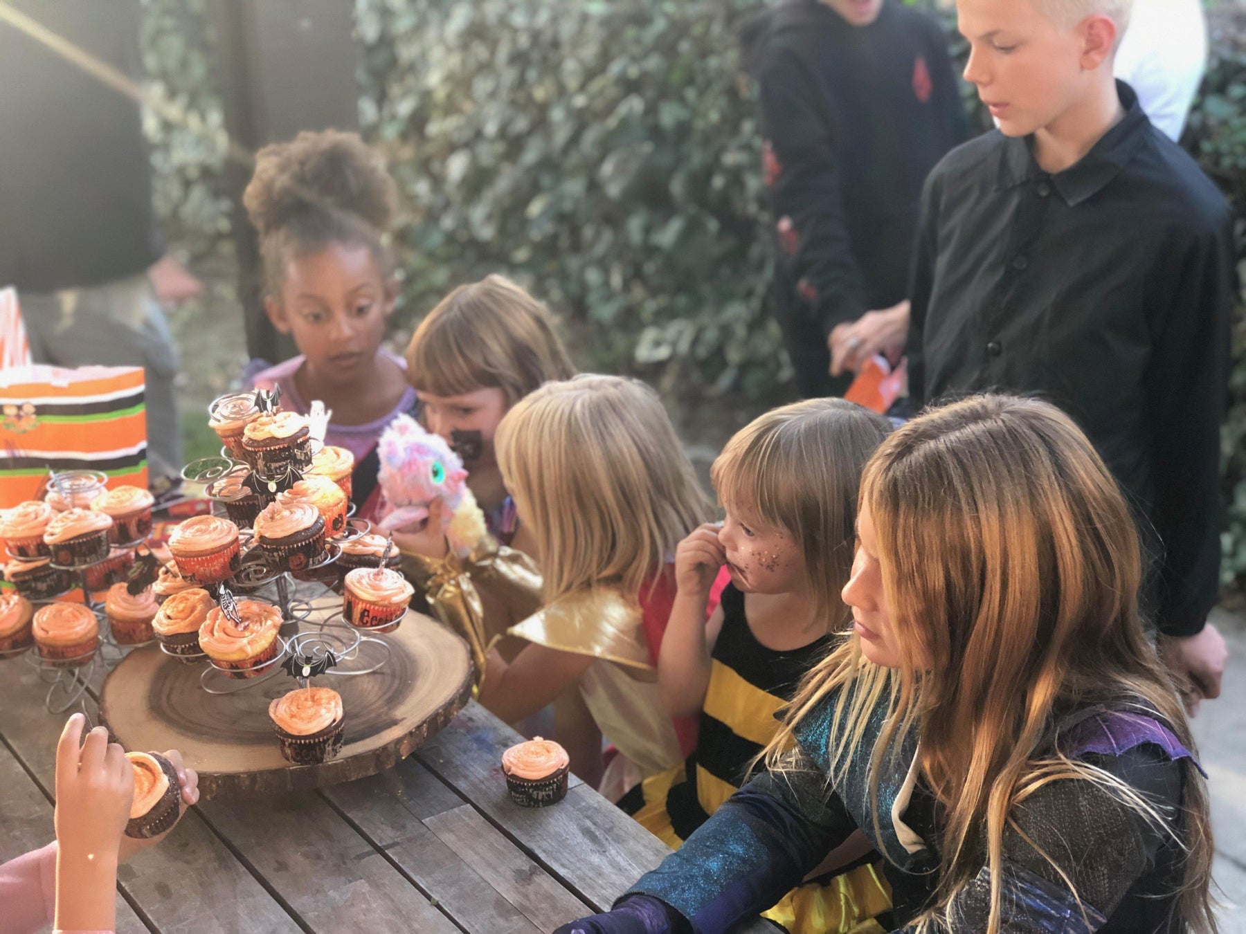 Kids in Halloween costumes gather around a rustic wooden cupcake stand with orange-frosted treats.