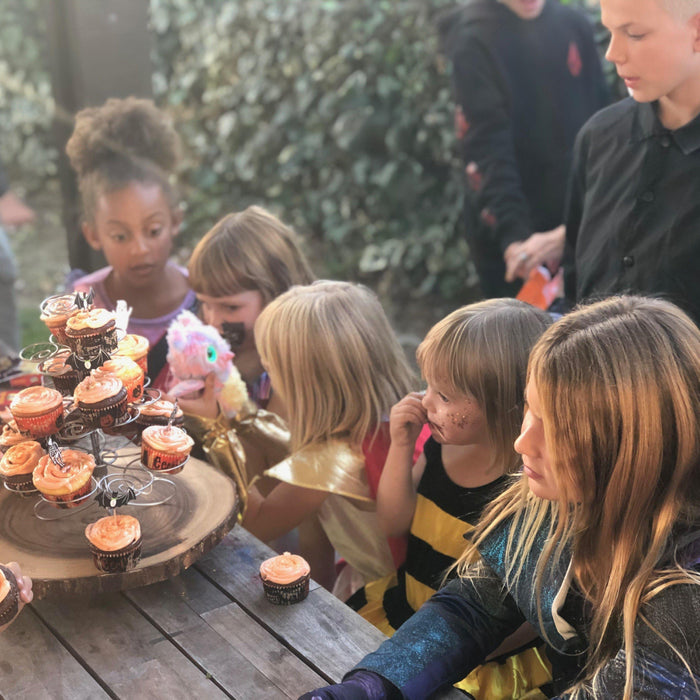 Kids in Halloween costumes gather around a rustic wooden cupcake stand with orange-frosted treats.