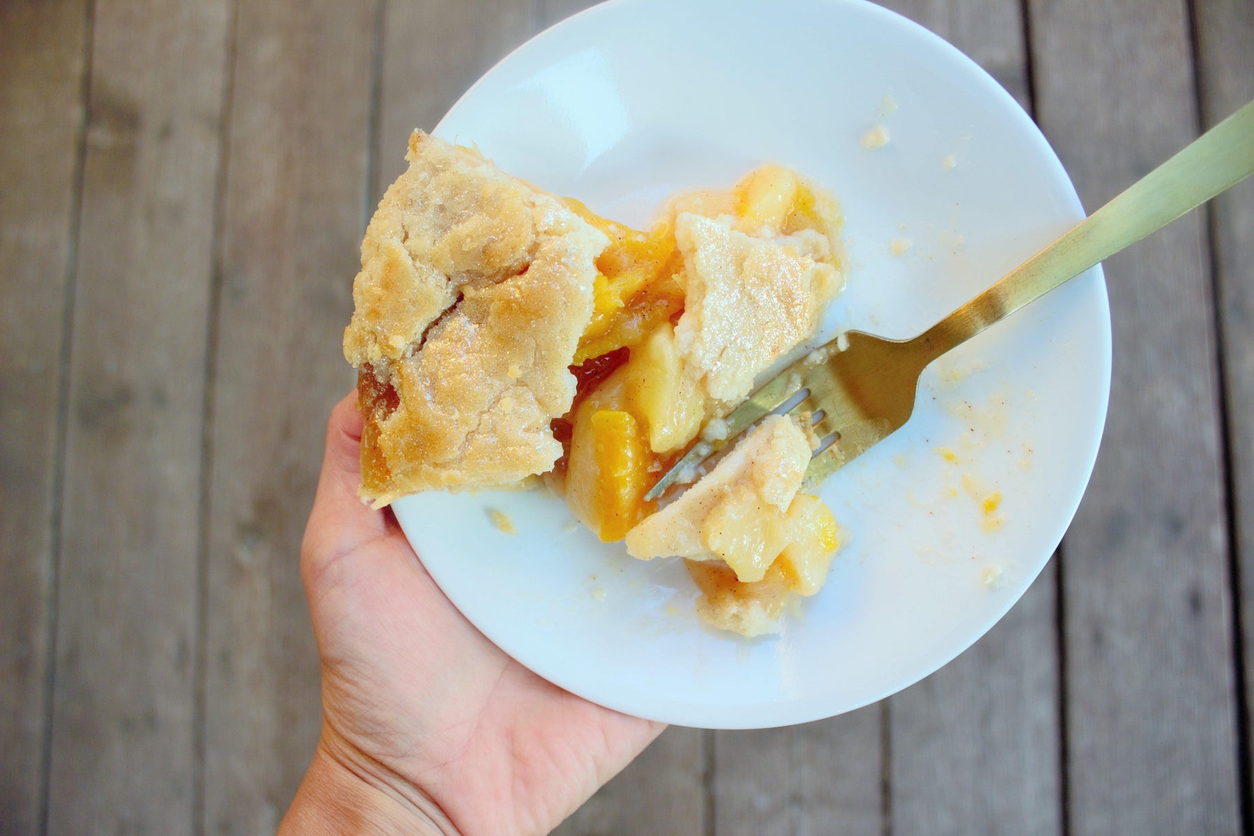 Hand holding a white plate with a slice of peach and apple pie, a golden fork taking a bite.
