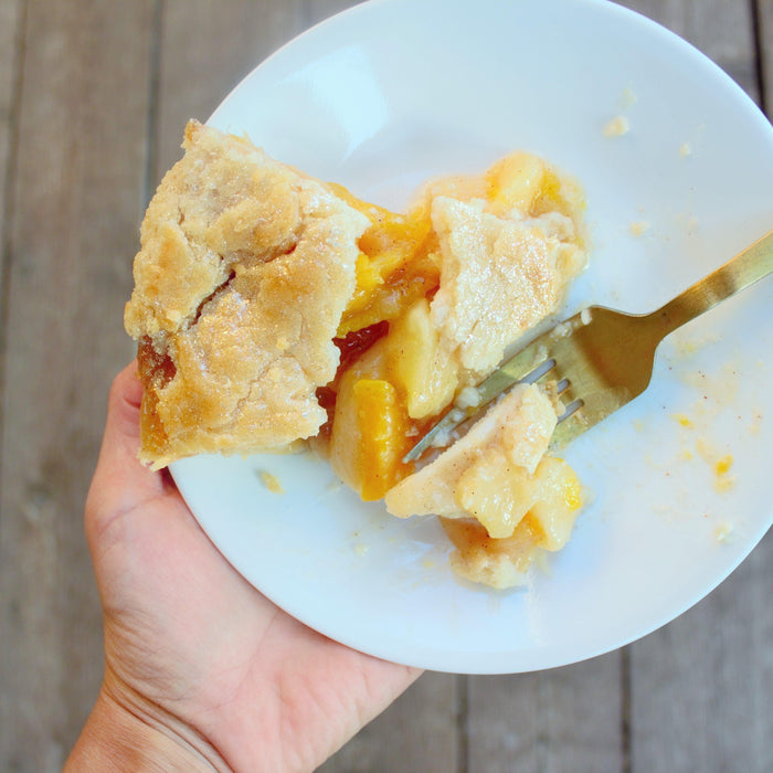 Hand holding a white plate with a slice of peach and apple pie, a golden fork taking a bite.