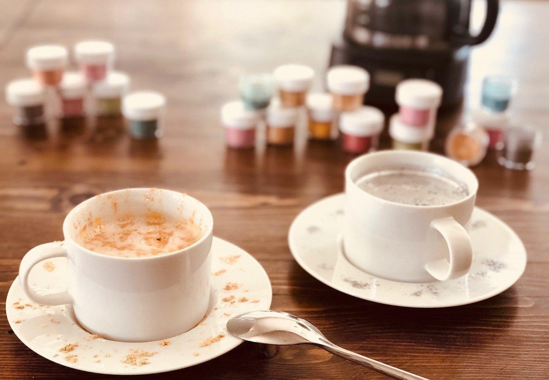 Two white mugs of coffee on a wooden table, with colorful spice jars in the background.
