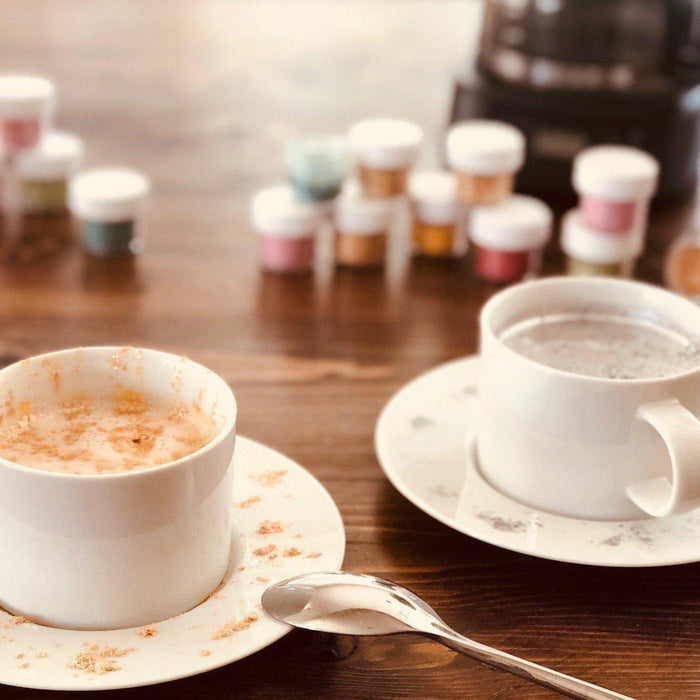 Two white mugs of coffee on a wooden table, with colorful spice jars in the background.