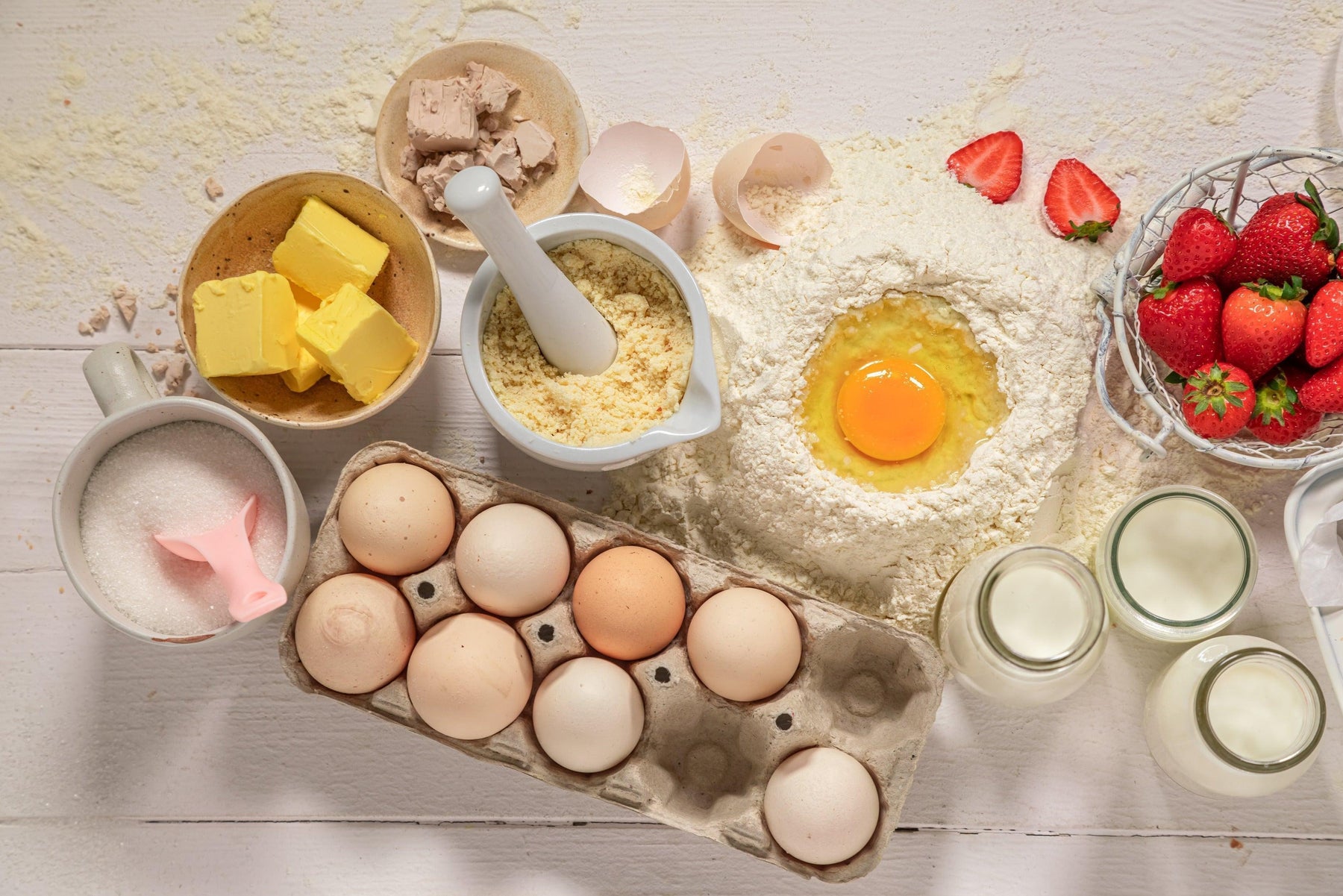 Baking ingredients: flour, eggs, butter, sugar, yeast, milk, and fresh strawberries on a white wooden table.