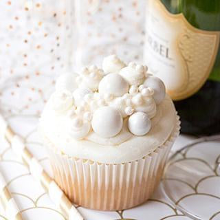 Elegant white cupcake with pearl decorations, gold striped straws, and a champagne bottle.
