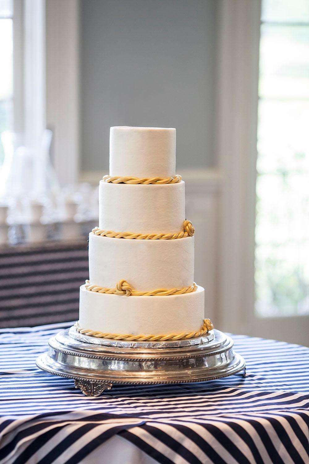 Four-tier white wedding cake with gold rope and knot details on a silver stand, on a blue and white striped tablecloth.