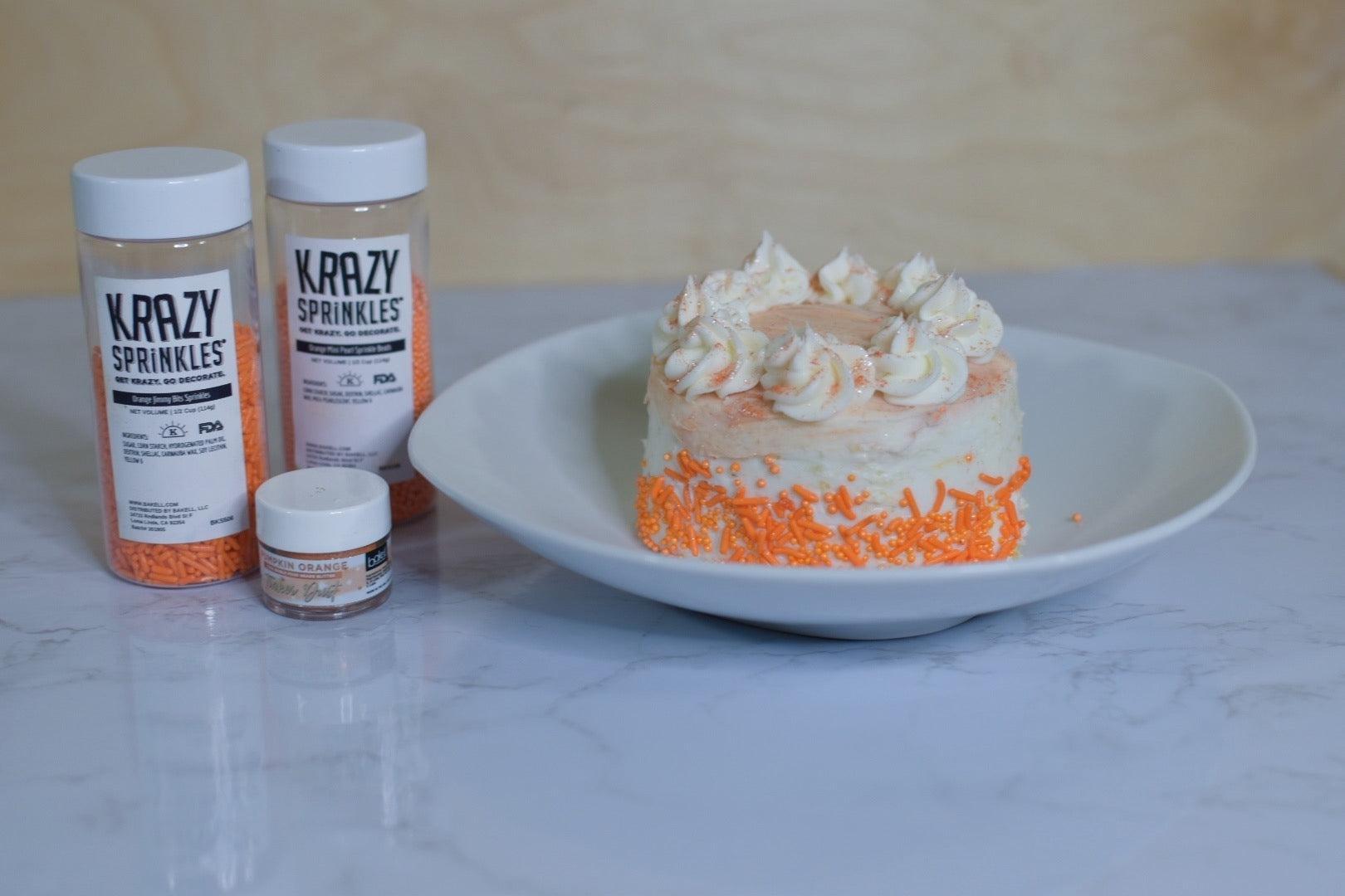 Mini orange cake with white frosting and orange sprinkles, next to Krazy Sprinkles bottles.