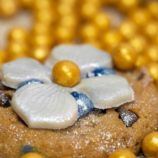 Sparkling chocolate chip cookie with white flower icing, blue glitter, and gold pearl candies.