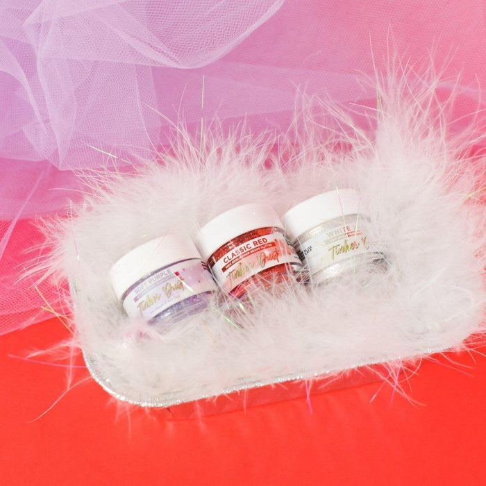 Three small jars of purple, red, and white glitter on a fluffy white tray, against a red background.