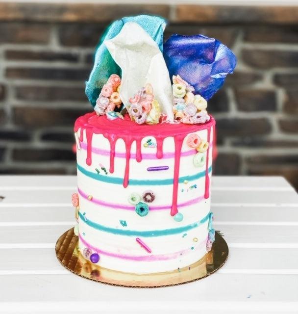 White cake with pink and blue stripes, pink drip icing, and colorful cereal and candy toppings.