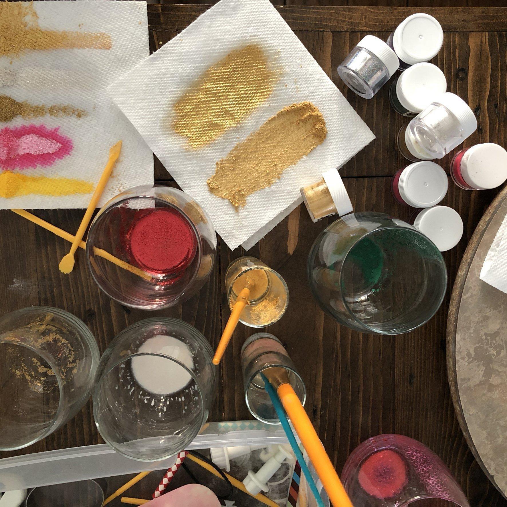 Crafting table with gold and colorful glitter, brushes, and glasses for DIY projects.