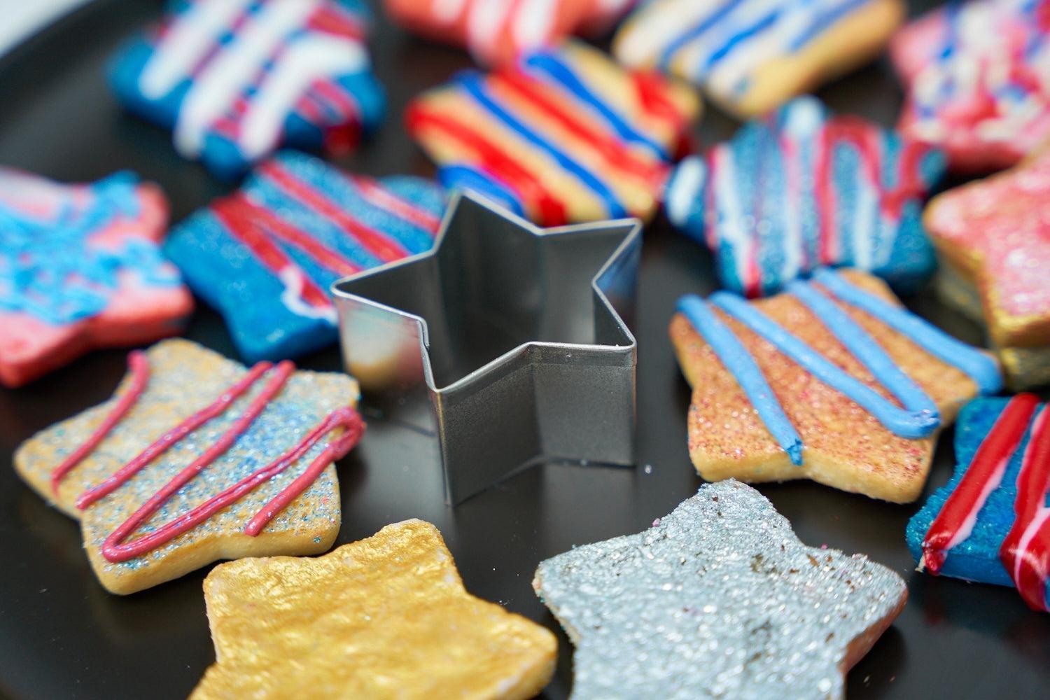 Star-shaped sugar cookies with red, white, and blue icing and sprinkles, surrounding a metal cookie cutter.