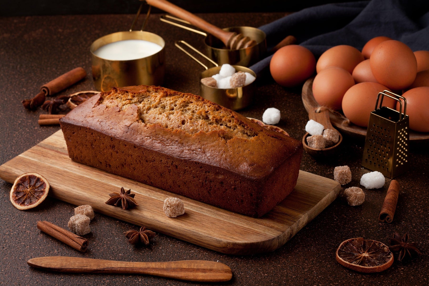 Freshly baked banana bread loaf on a wooden board, surrounded by eggs, milk, sugar, honey, and spices.