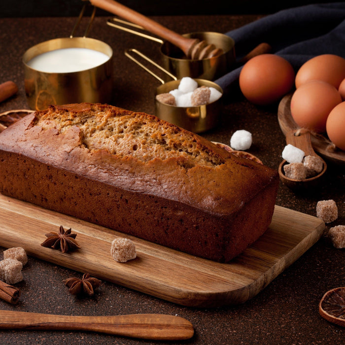 Freshly baked banana bread loaf on a wooden board, surrounded by eggs, milk, sugar, honey, and spices.