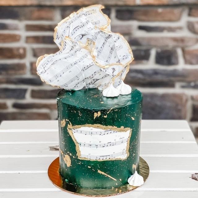 Elegant dark green cake with gold splatters, decorated with edible sheet music and a large musical note topper.