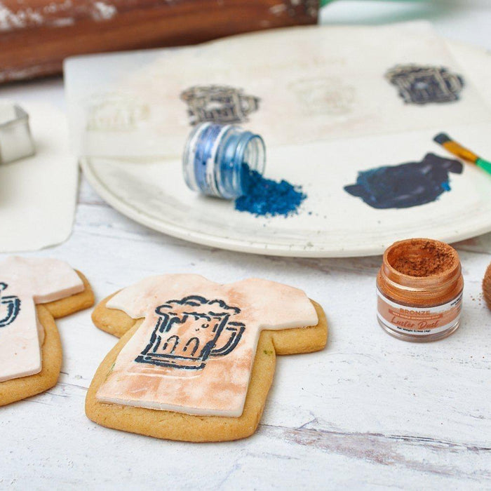Decorating T-shirt shaped cookies with beer mug designs, edible bronze luster dust, and blue glitter.