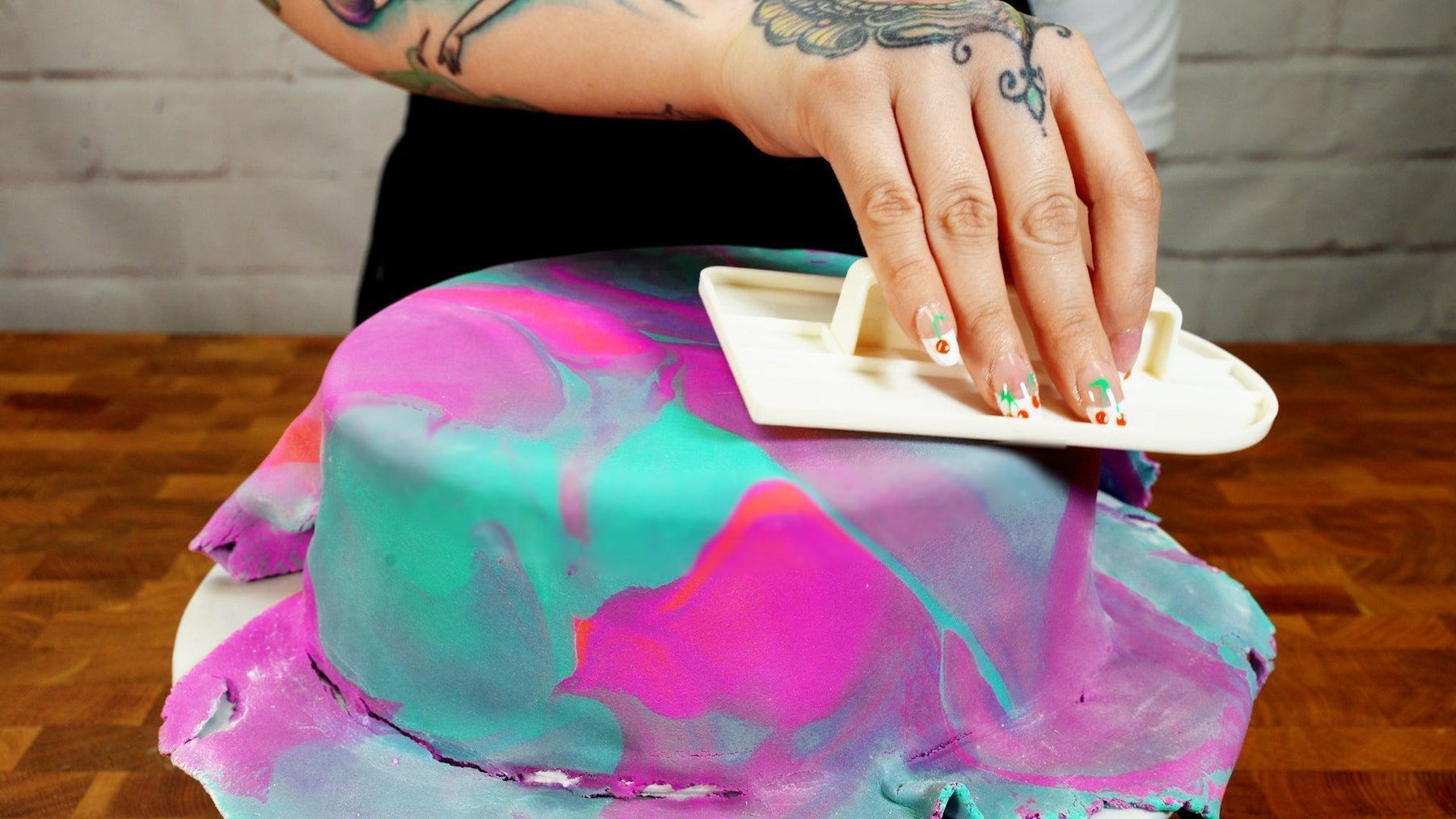 Hand smoothing vibrant pink and teal marbled fondant on a cake with a white tool.