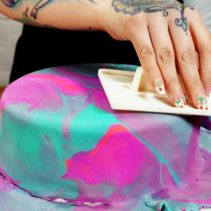 Hand smoothing vibrant pink and teal marbled fondant on a cake with a white tool.