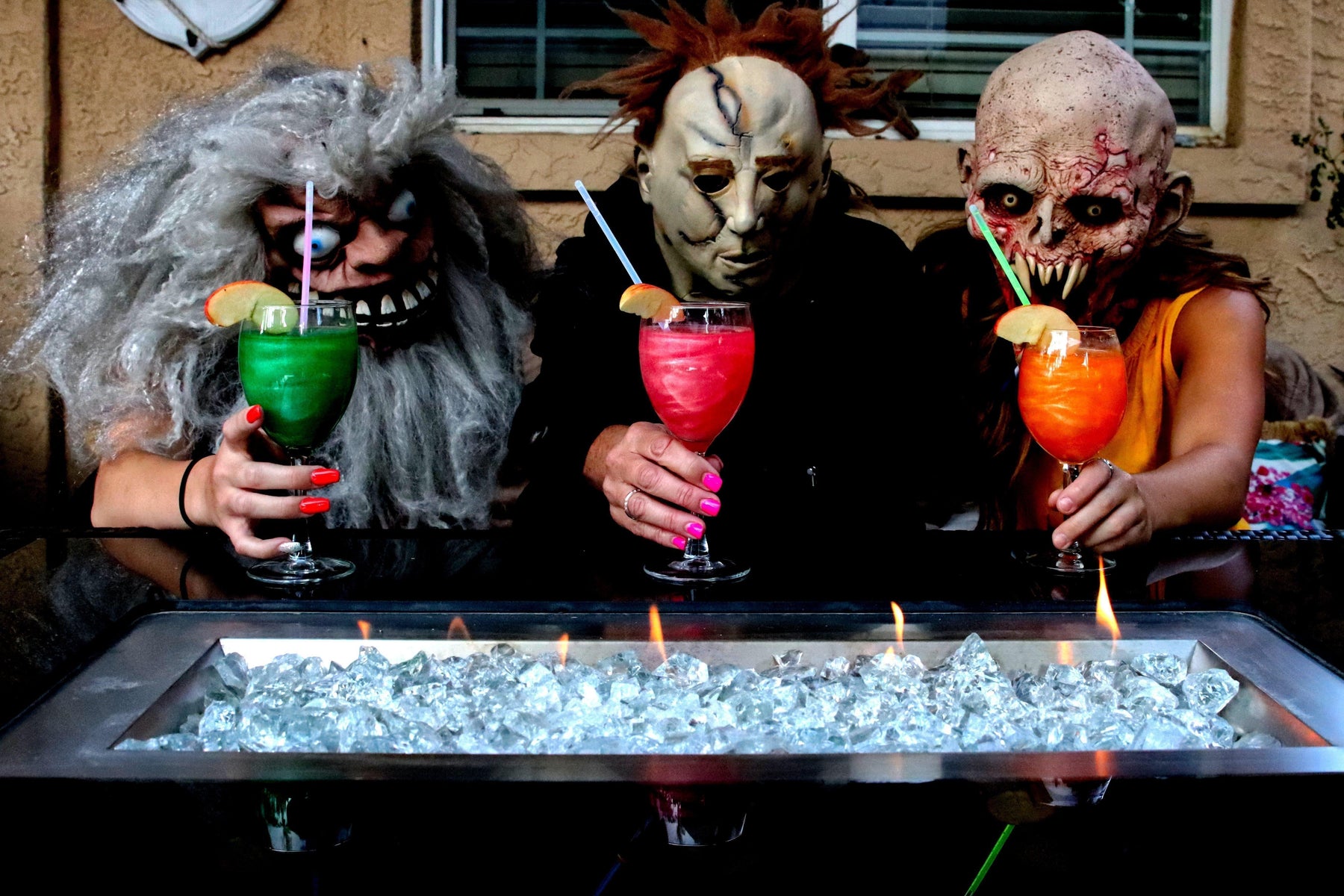 Three people in scary Halloween masks hold colorful cocktails over a fire pit.