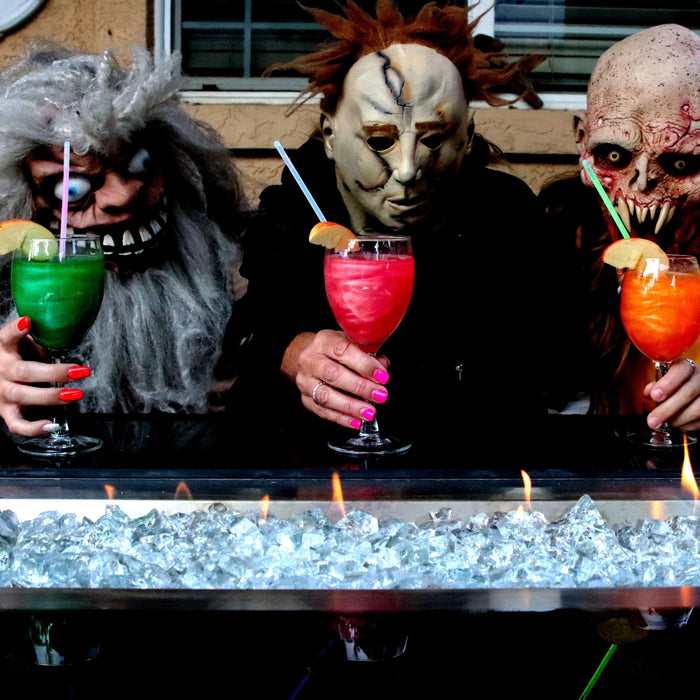 Three people in scary Halloween masks hold colorful cocktails over a fire pit.