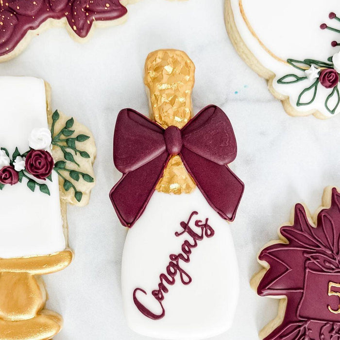Elegant champagne bottle cookie with a burgundy bow and "Congrats" in script, surrounded by floral-decorated cookies.