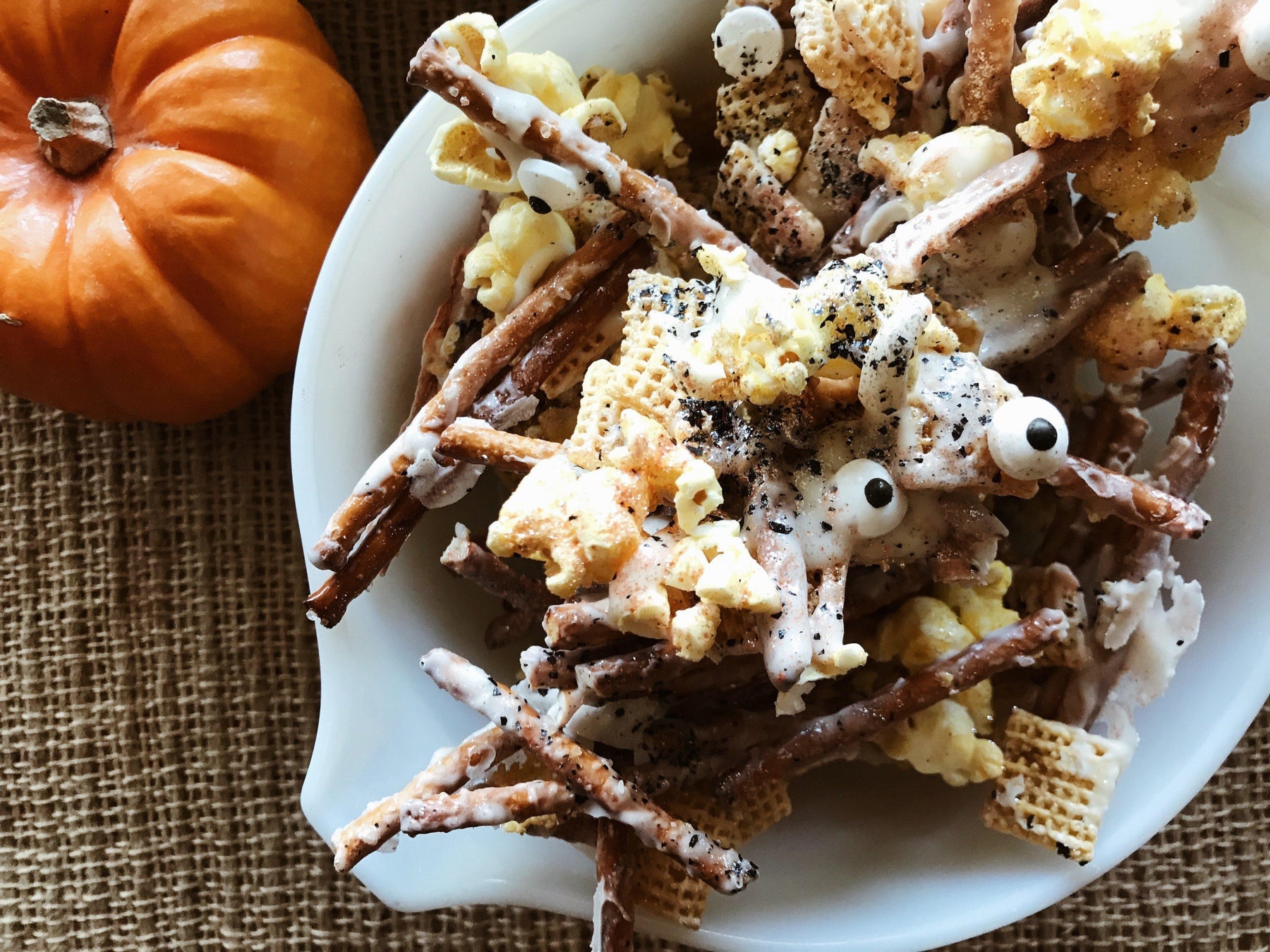 White bowl of Halloween monster munch mix with pretzels, popcorn, cereal, candy eyes, and a pumpkin.