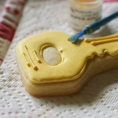 Decorating a key-shaped sugar cookie with yellow icing and edible gold paint using a small blue brush.