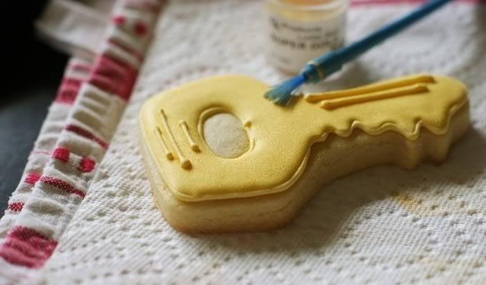 Decorating a key-shaped sugar cookie with yellow icing and edible gold paint using a small blue brush.
