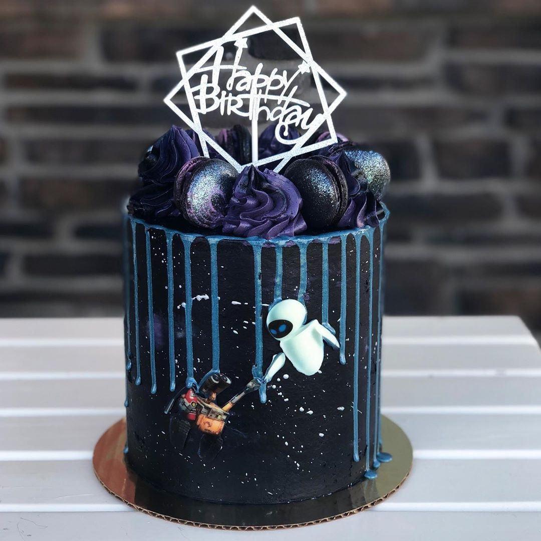Dark blue birthday cake with Wall-E and EVE characters, purple frosting, macarons, and blue drip icing.