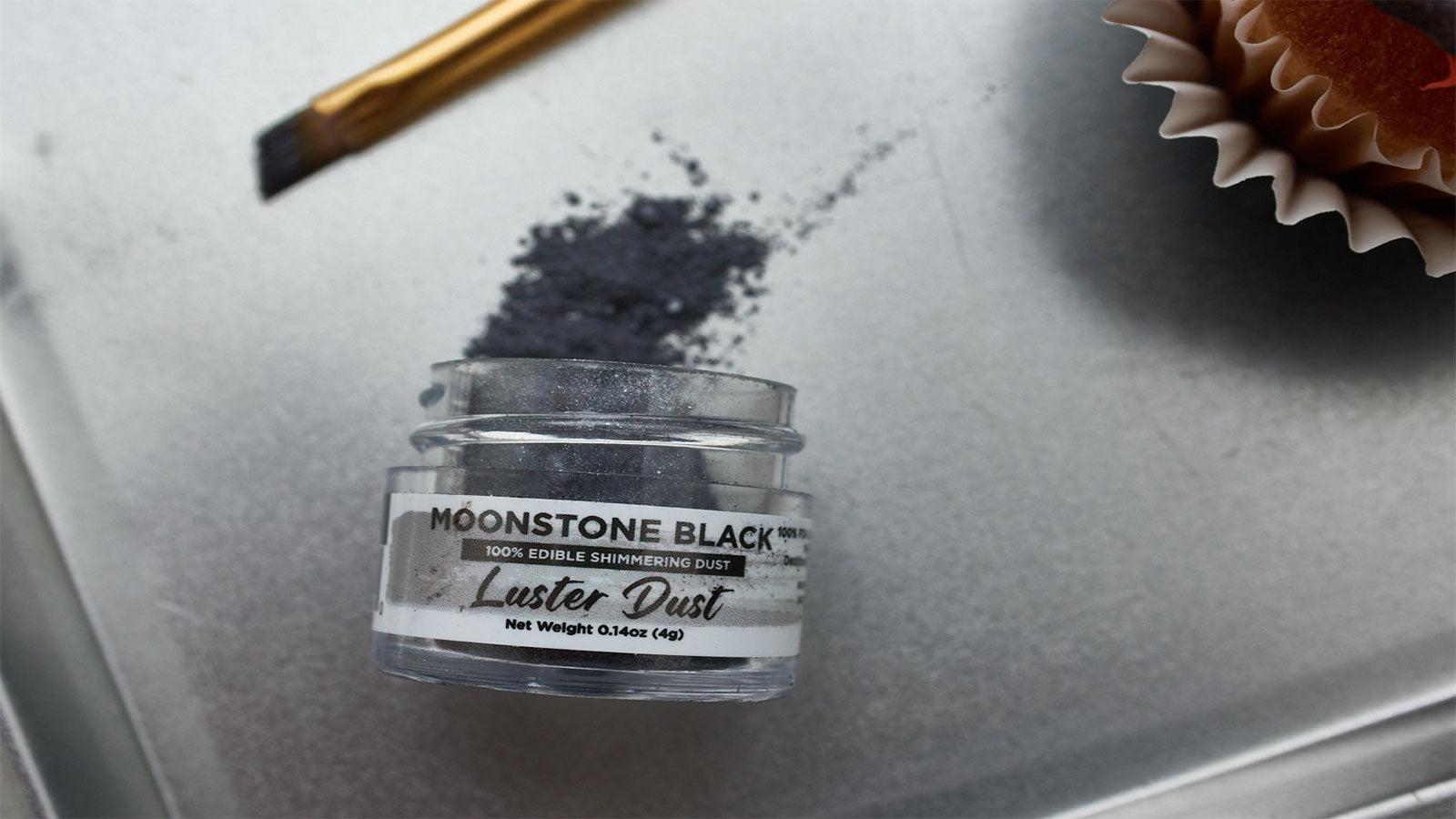 Moonstone Black edible luster dust in a clear jar, with a brush and cupcake liner on a silver tray.