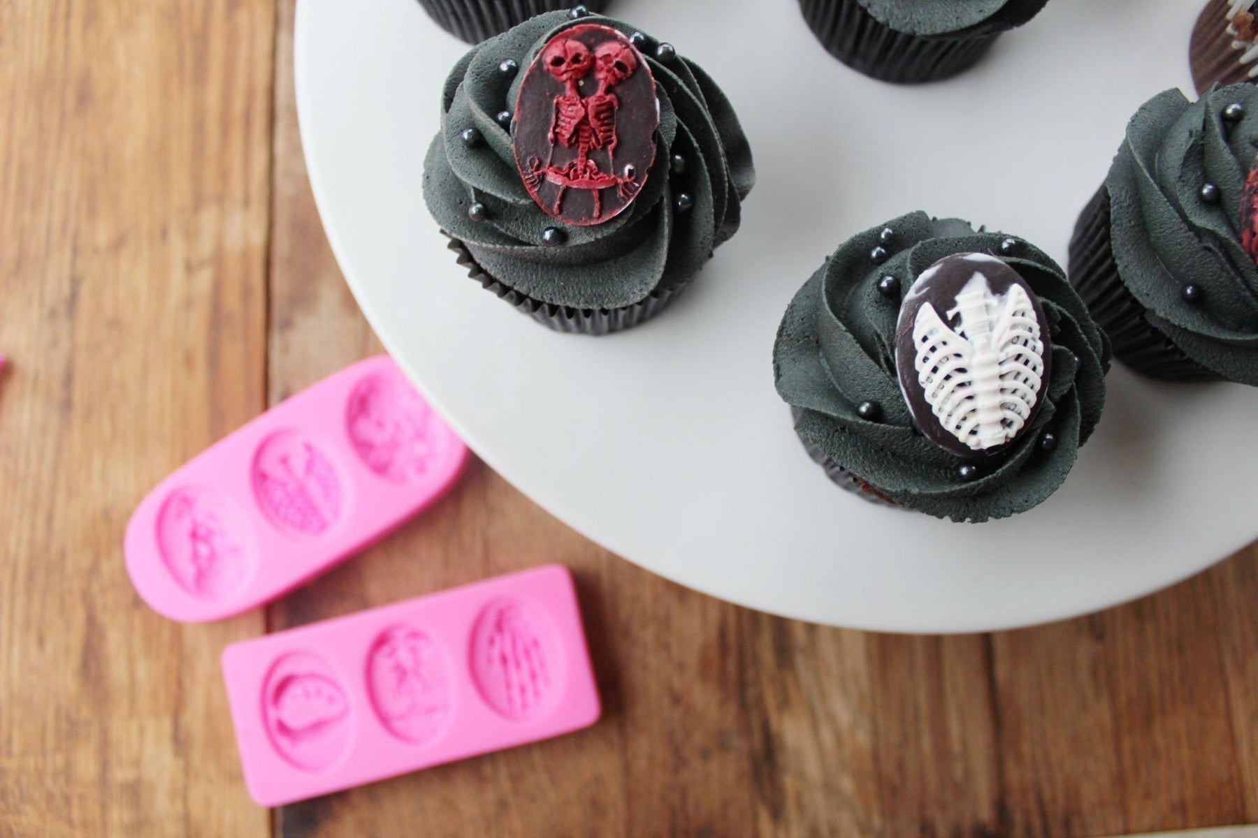 Dark Halloween cupcakes with skeleton and skull cameos, next to pink silicone molds.