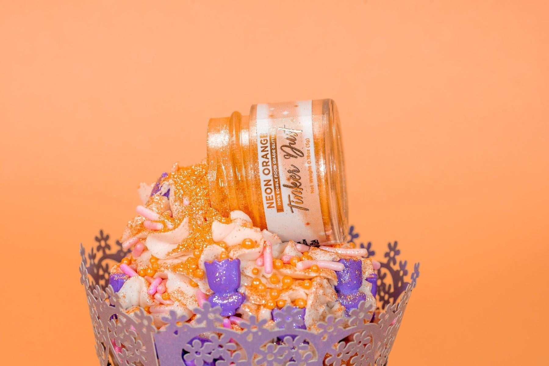 Orange Tinker Dust edible glitter spilling from a jar onto a frosted cupcake with purple and pink sprinkles.