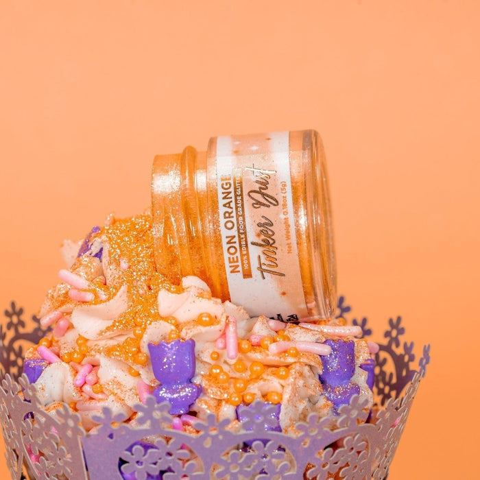 Orange Tinker Dust edible glitter spilling from a jar onto a frosted cupcake with purple and pink sprinkles.