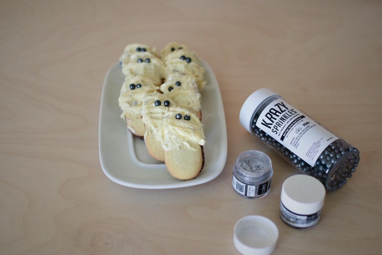 Mummy-shaped cookies with white frosting and black sprinkles, next to Krazy Sprinkles and glitter.