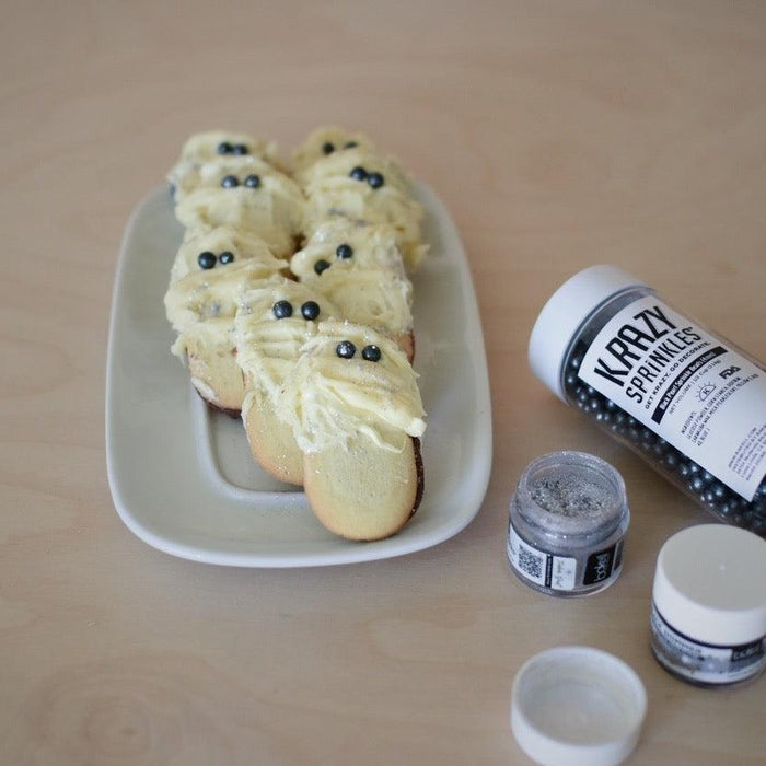 Mummy-shaped cookies with white frosting and black sprinkles, next to Krazy Sprinkles and glitter.