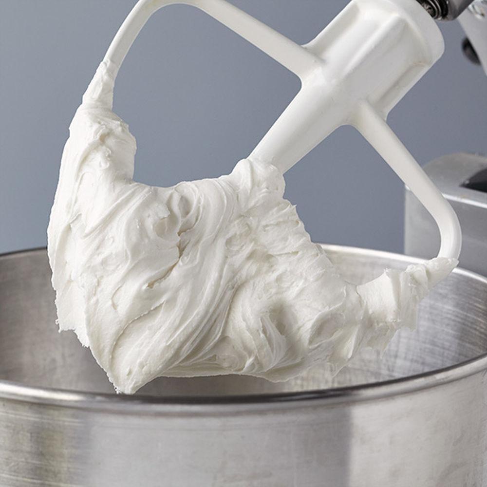 White paddle attachment covered in fluffy, creamy frosting over a stainless steel mixing bowl.