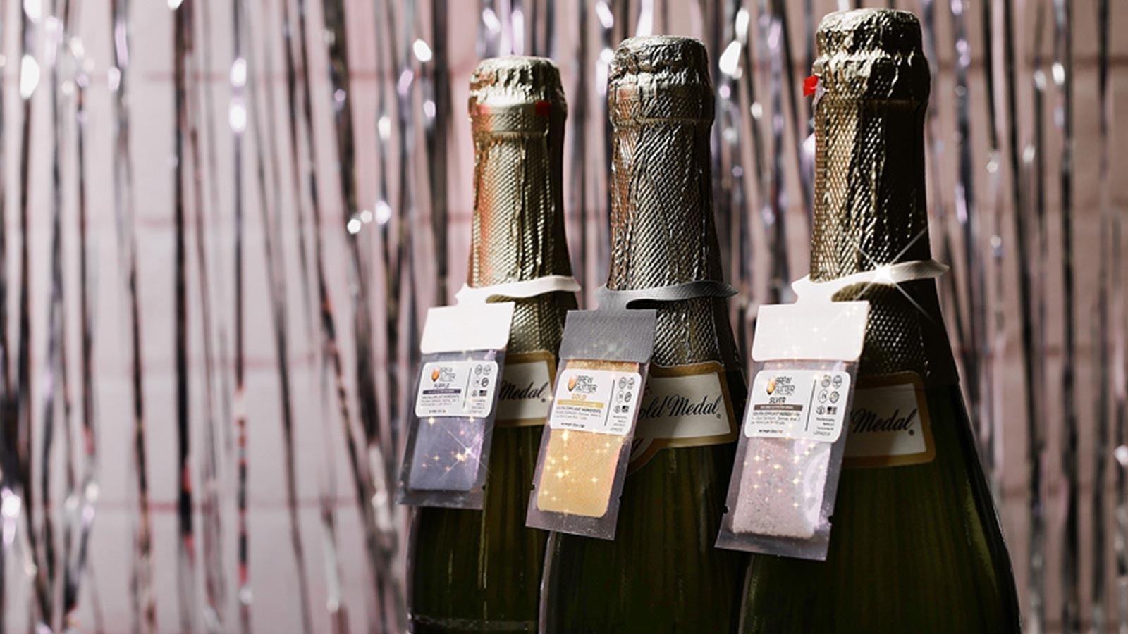 Three champagne bottles with edible glitter packets for festive drinks against a silver tinsel background.