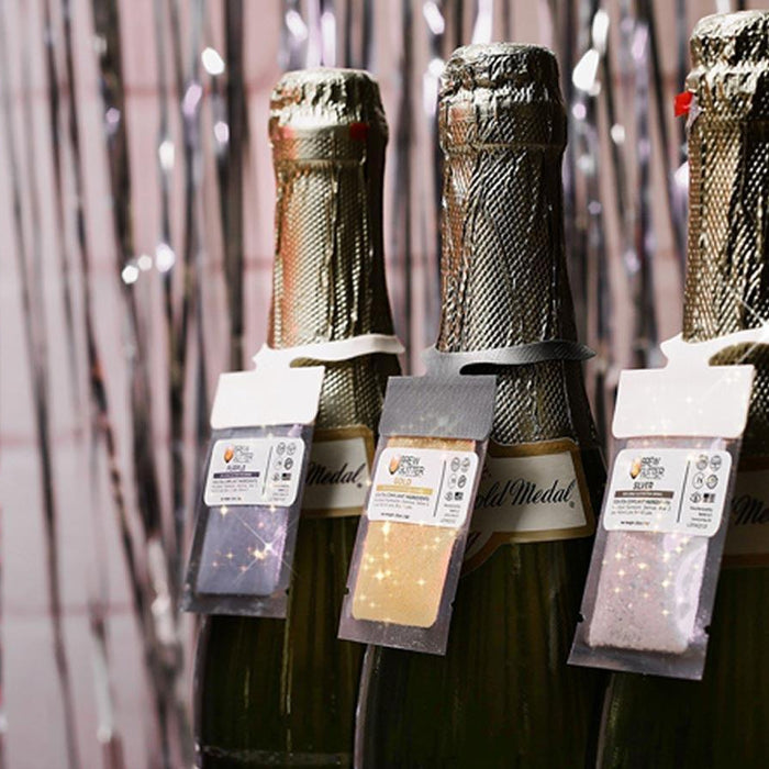 Three champagne bottles with edible glitter packets for festive drinks against a silver tinsel background.