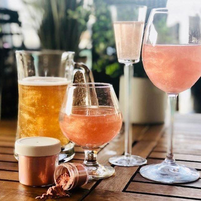 Shimmering pink and gold drinks in various glasses, with edible glitter pots on a wooden table.