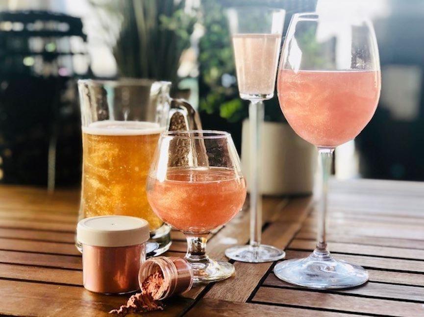 Shimmering pink and gold drinks in various glasses, with edible glitter pots on a wooden table.