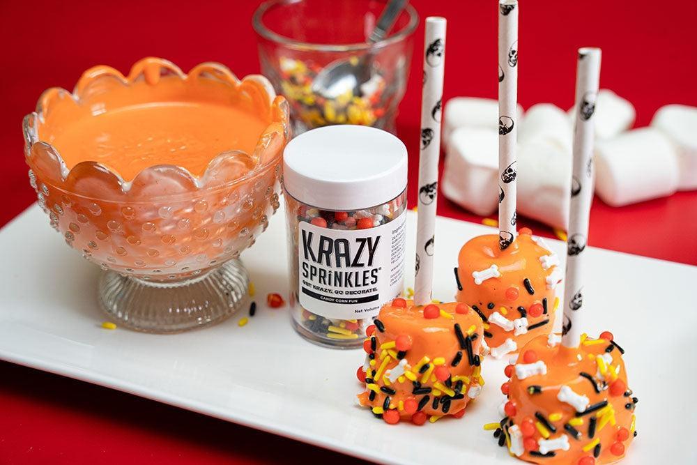 Orange marshmallow pops with skull straws and bone sprinkles, alongside a bowl of orange dip and Krazy Sprinkles.