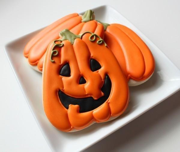 Bright orange pumpkin-shaped Halloween cookies with black jack-o'-lantern faces and green vine details.
