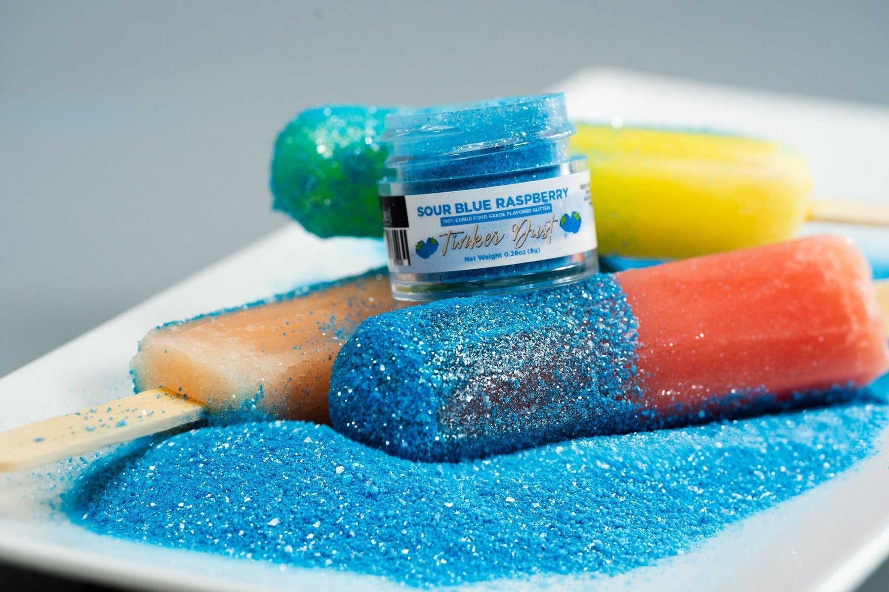 Blue raspberry edible glitter on popsicles with a jar of "Sour Blue Raspberry Tinker Dust.