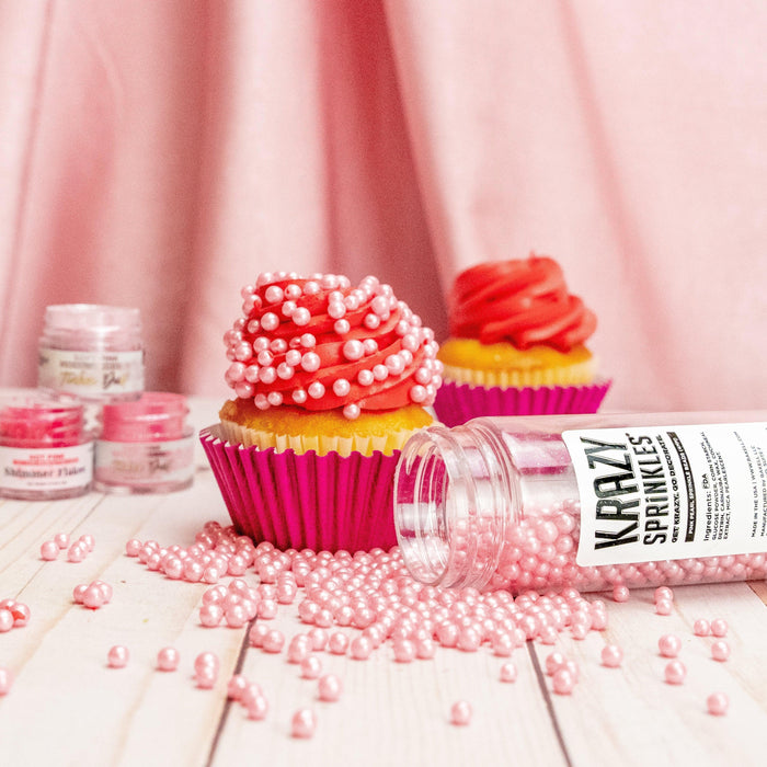 Pink Krazy Sprinkles spilled from a bottle next to two cupcakes on a wooden table.