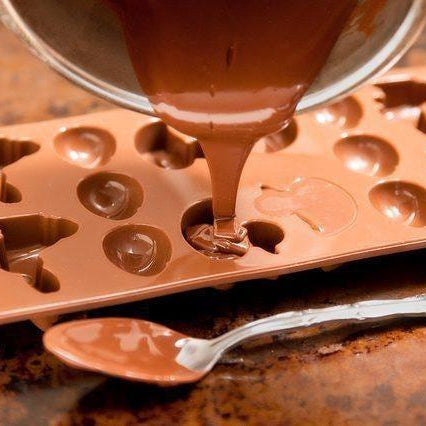 Melted chocolate pouring from a saucepan into a brown silicone candy mold with various shapes.