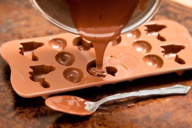 Melted chocolate pouring from a saucepan into a brown silicone candy mold with various shapes.