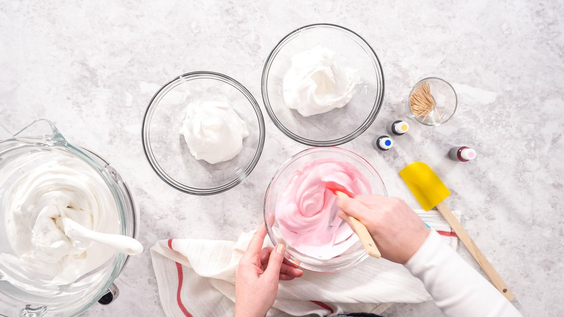 Hands mixing pink frosting in a glass bowl, with white frosting and food coloring nearby.