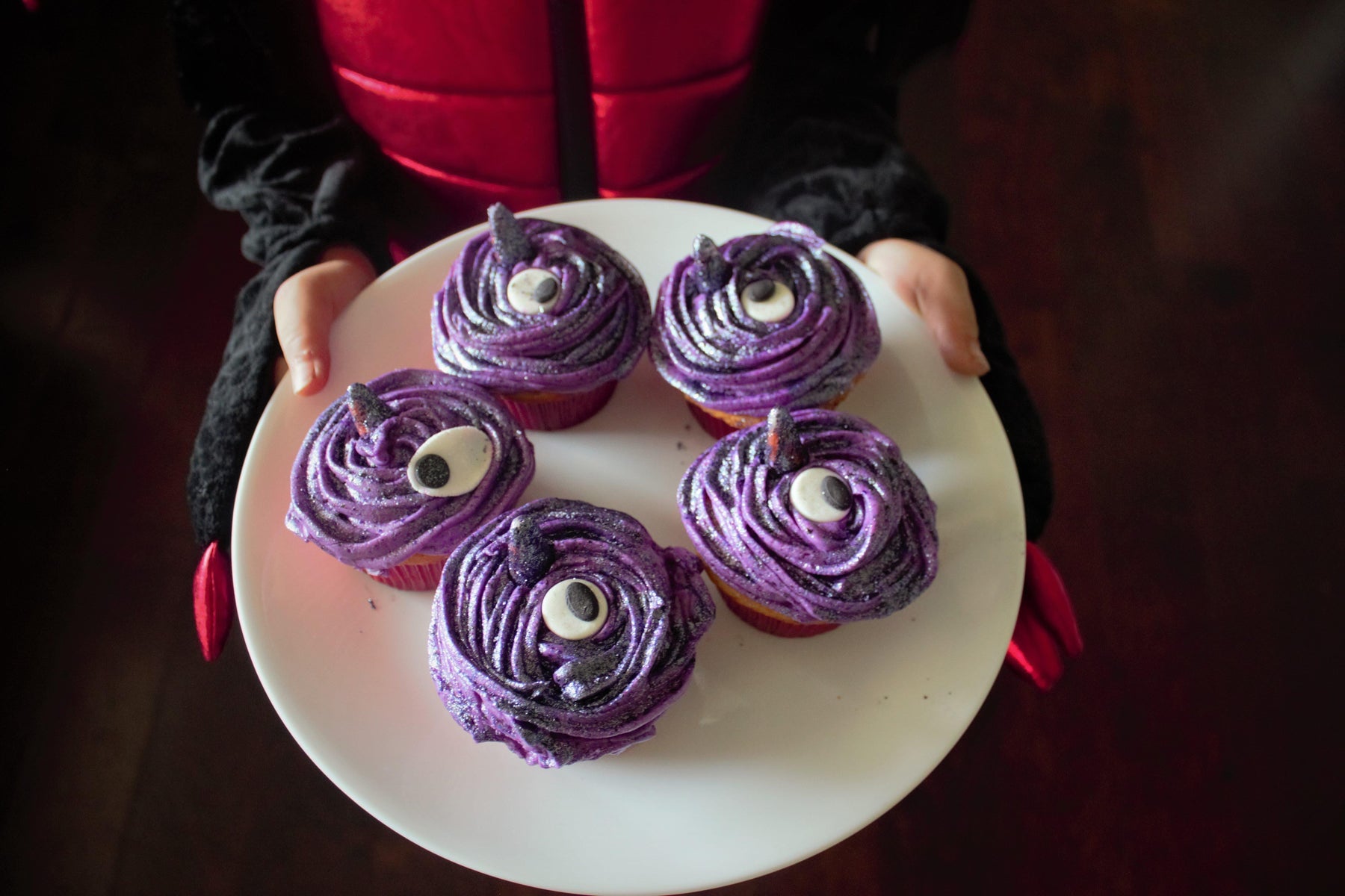 Child in a red and black costume holds a white plate with five purple monster cupcakes with googly eyes.