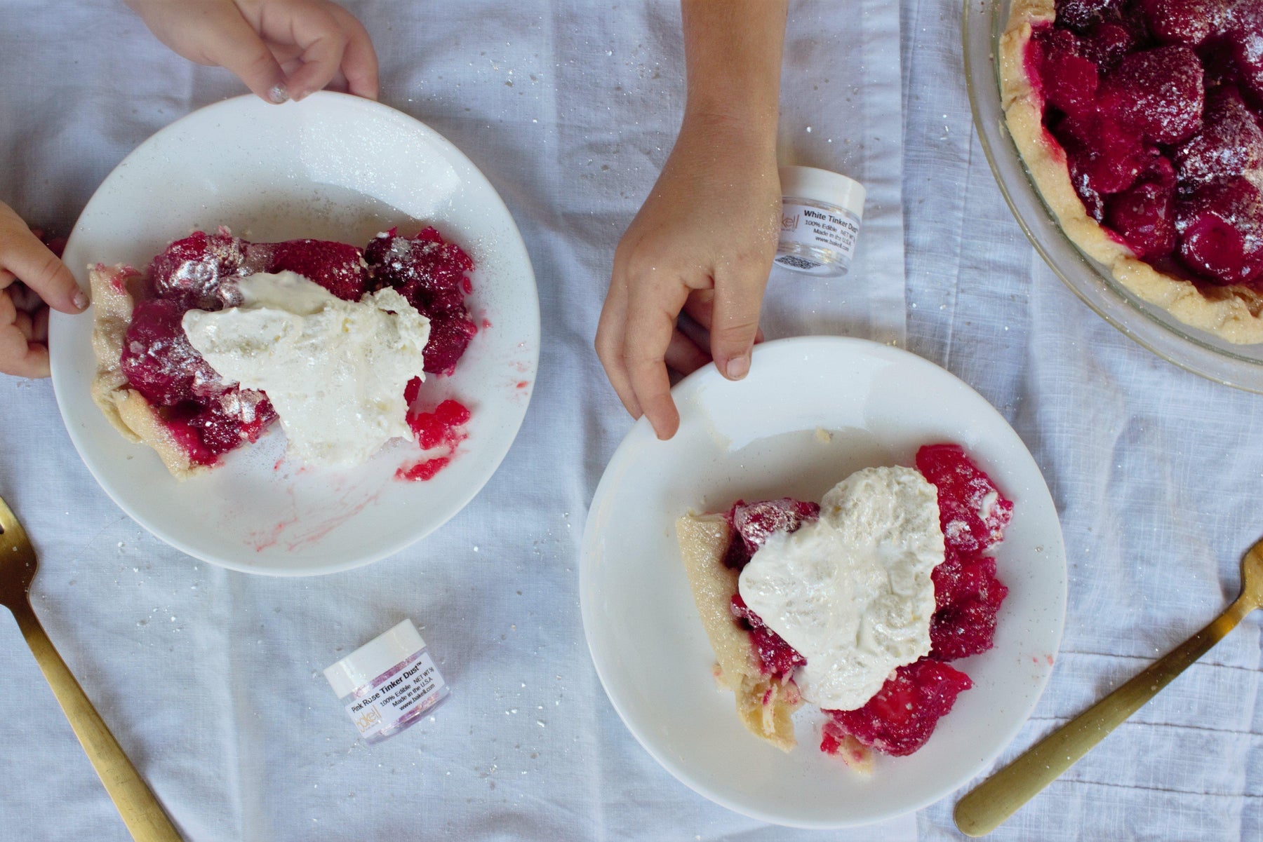 Two hands serving slices of vibrant red berry pie topped with whipped cream on white plates.